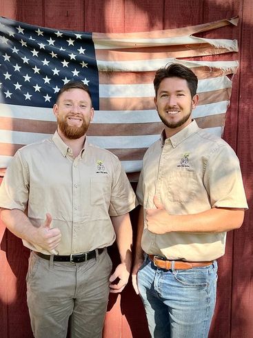 Two men in beige shirts standing before a faded American flag backdrop, smiling and giving thumbs up