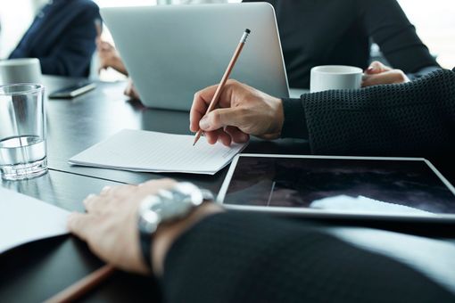 Un uomo sta scrivendo su un pezzo di carta mentre è seduto a un tavolo con un computer portatile e un tablet.