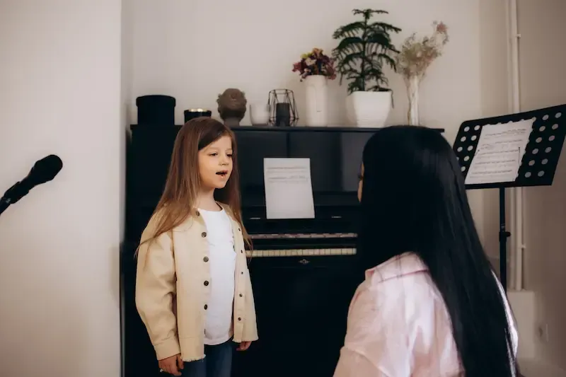 A child stands in a music room singing while facing an instructor, with a piano and a music stand in the background.