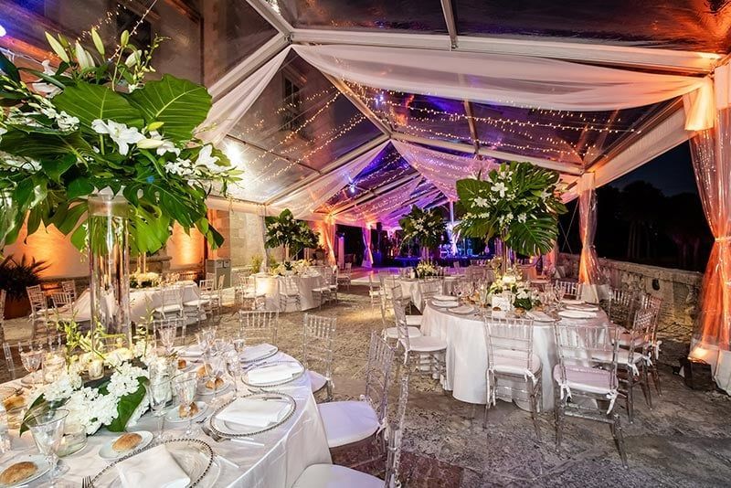 Wedding reception setup inside a clear tent, round tables with white linens, floral centerpieces.