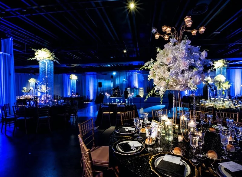 Elegant event space with round tables, floral centerpieces, blue lighting, and gold chairs.