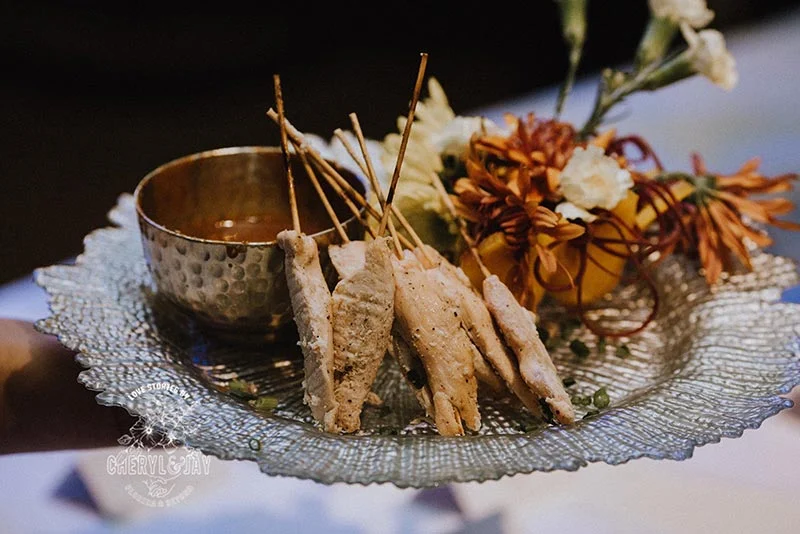 Chicken skewers with sauce, floral arrangement on decorative glass plate.
