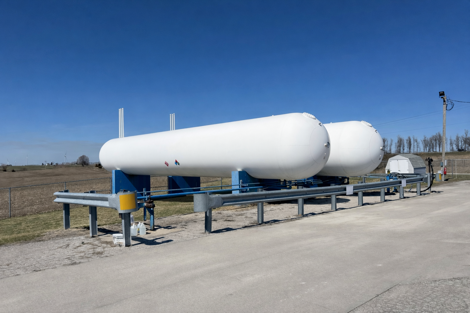 Two large, white horizontal propane storage tanks supported by blue metal stands in an open field under a blue sky.