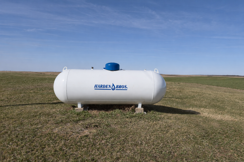 A white horizontal propane tank sits on concrete supports in a grassy field under a clear blue sky.
