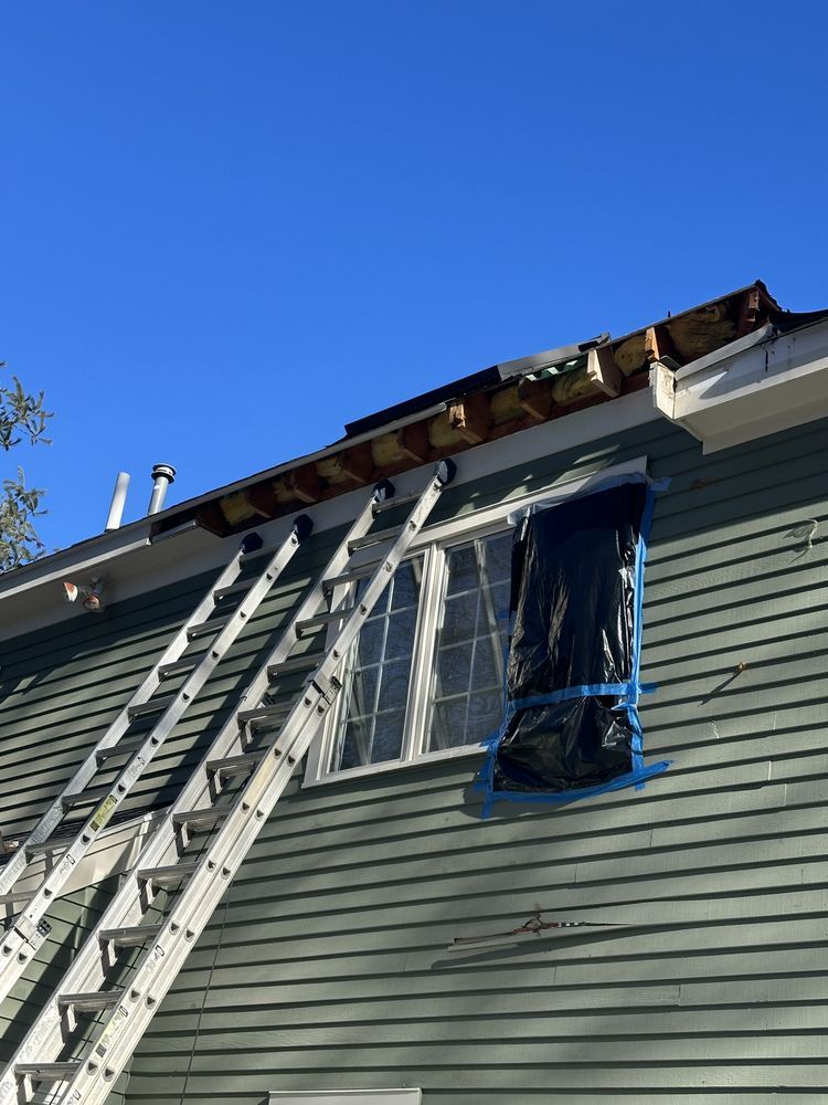 Ladders against a house with roof damage and a blue-taped window under a clear sky