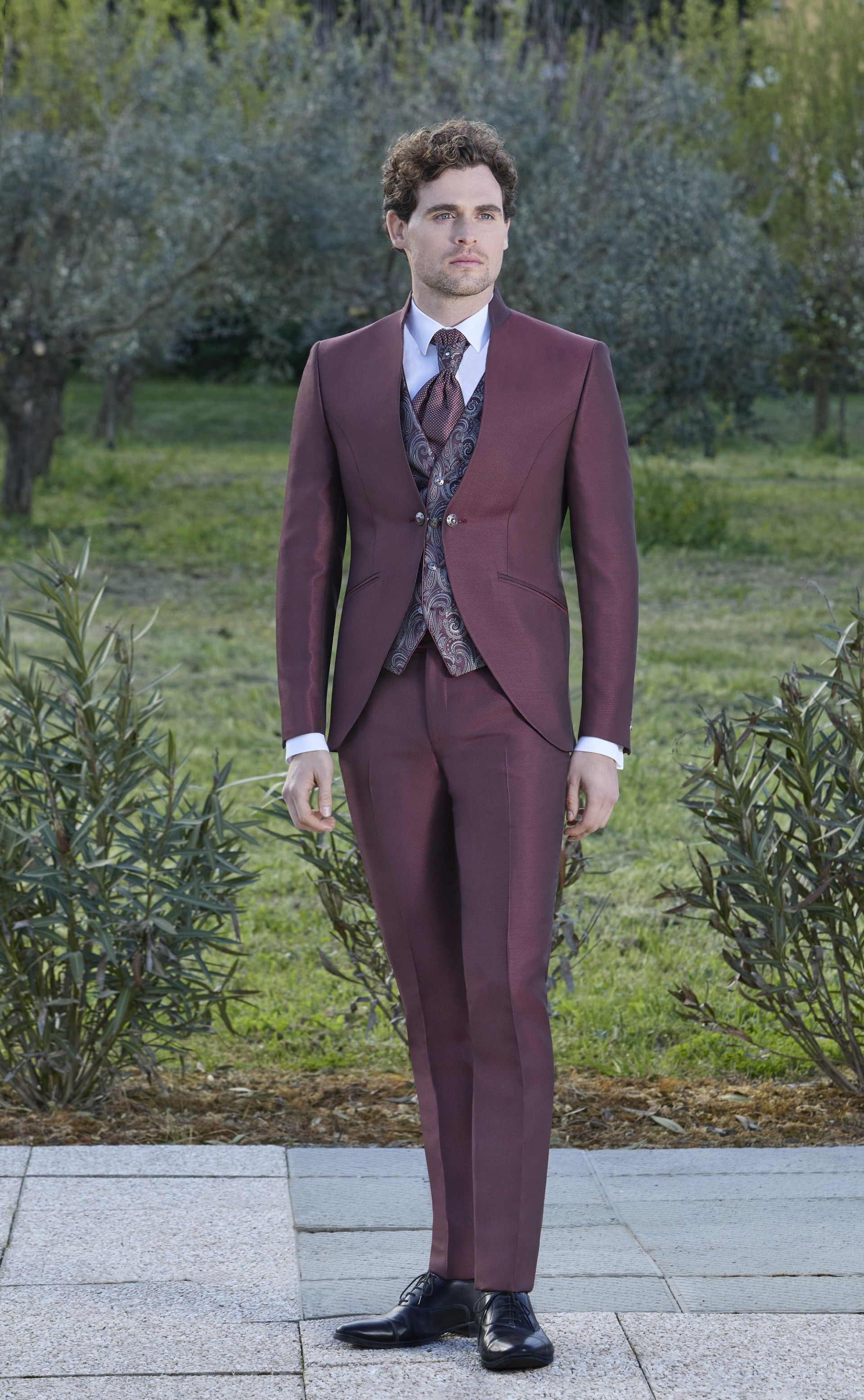 Un uomo in giacca e cravatta color bordeaux è in piedi sul marciapiede.