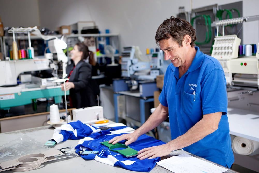 Man in Blue Shirt Examines Fabric on a Table in a Workshop — Workwise Clothing Forster in Forster, NSW