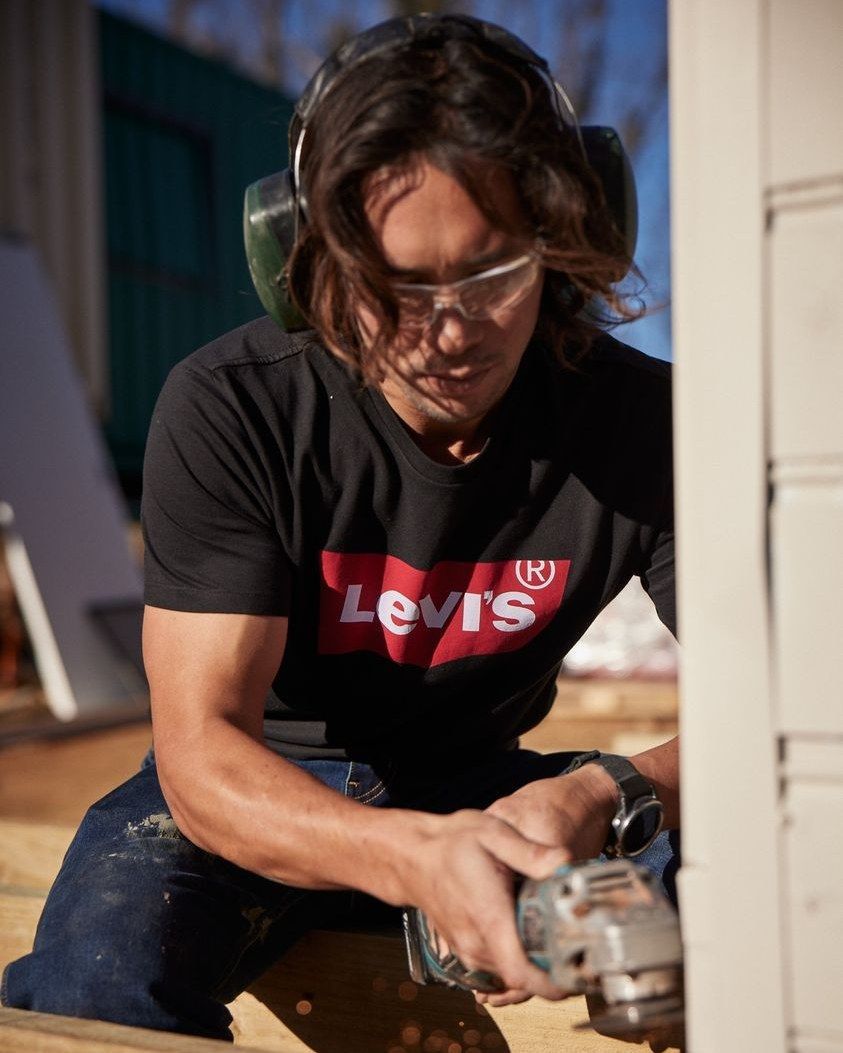 Man With Long Hair Wearing Safety Gear, Using a Grinder on Wood Outdoors — Workwise Clothing Forster in Wingham, NSW