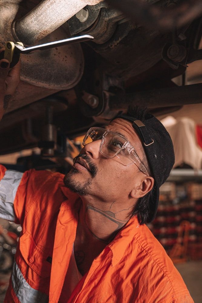 Mechanic Under a Car, Wearing Safety Glasses and Orange Overalls, Looking Up — Workwise Clothing Forster in Forster, NSW