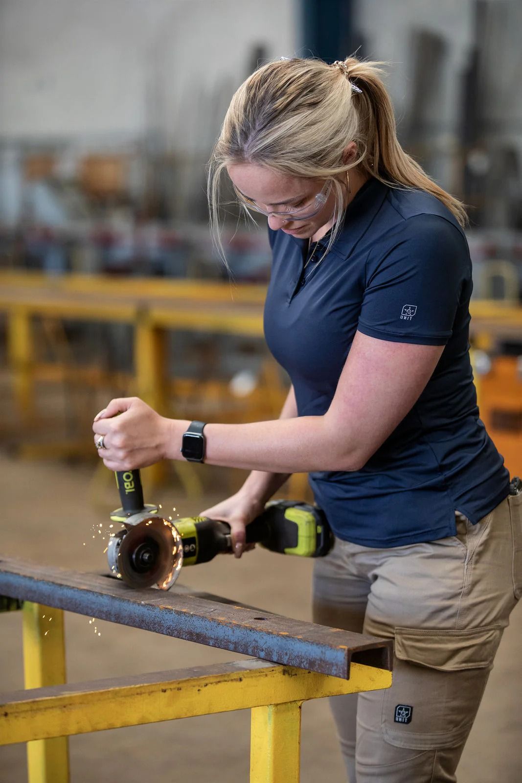 Woman in safety glasses grinds metal with a power tool in a workshop. Sparks fly — Workwise Clothing Forster in Forster, NSW