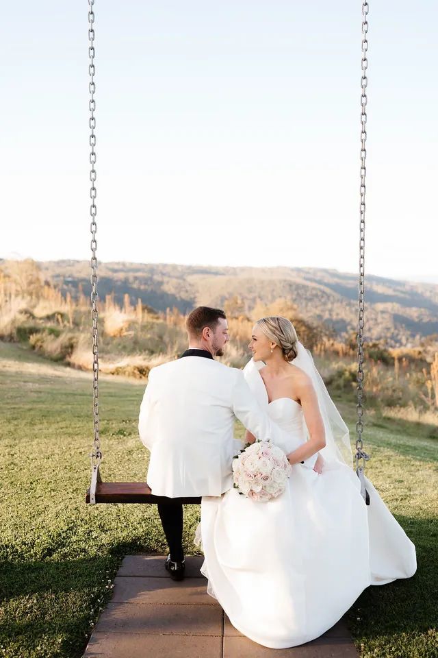 A bride and groom are sitting on a swing in a field.