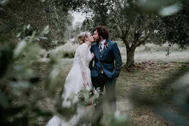 A bride and groom are kissing in a field surrounded by trees.