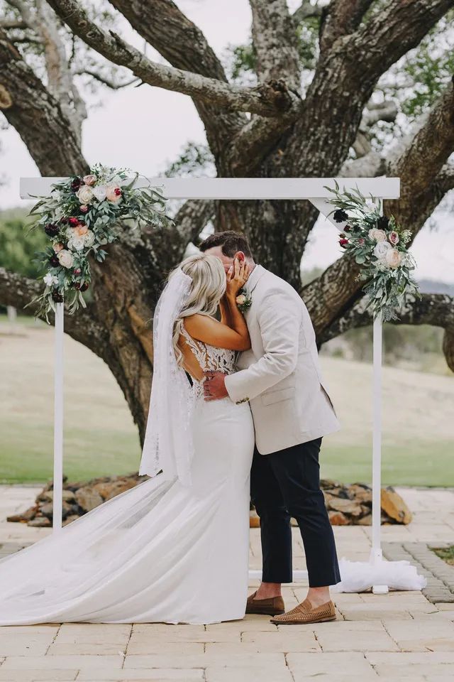 A bride and groom are kissing under a tree at their wedding ceremony.