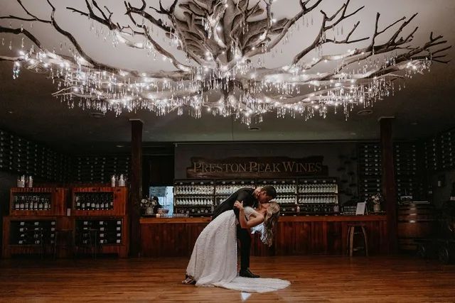 A bride and groom are dancing under a chandelier in a wine cellar.