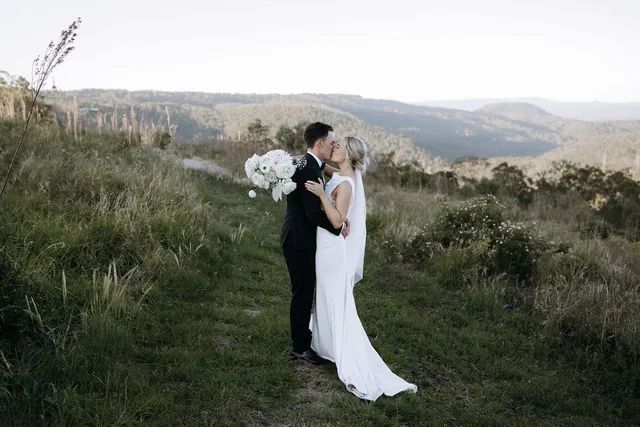 A bride and groom are kissing in a field with mountains in the background.