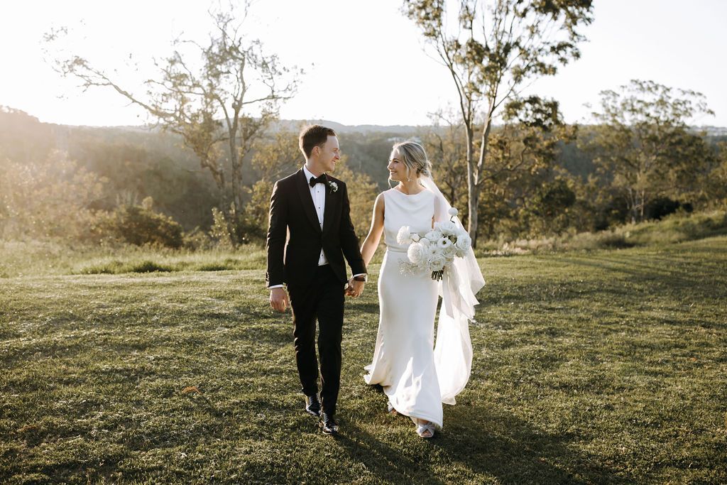 A bride and groom are walking in a field holding hands.