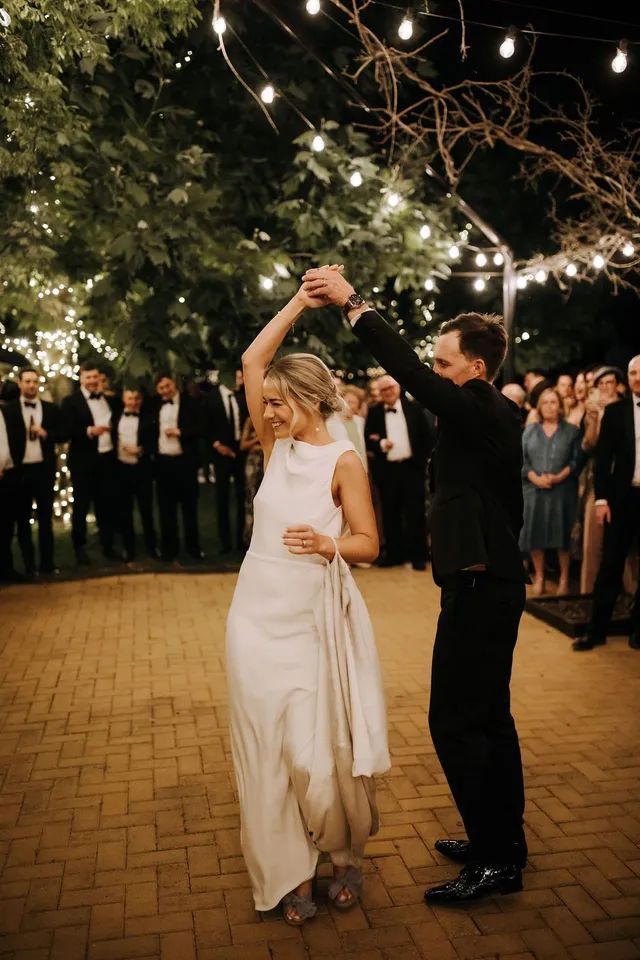 A bride and groom are dancing at their wedding reception under string lights.