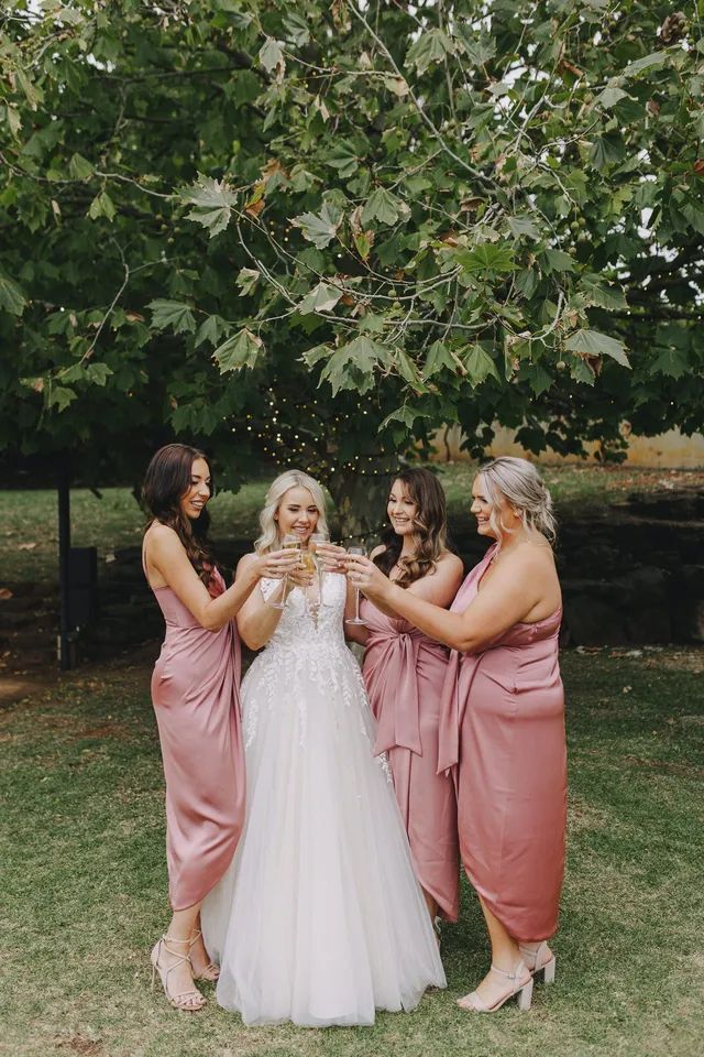 A bride and her bridesmaids are toasting with champagne in front of a tree.