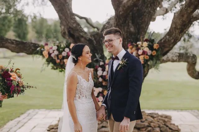 A bride and groom are holding hands in front of a tree.