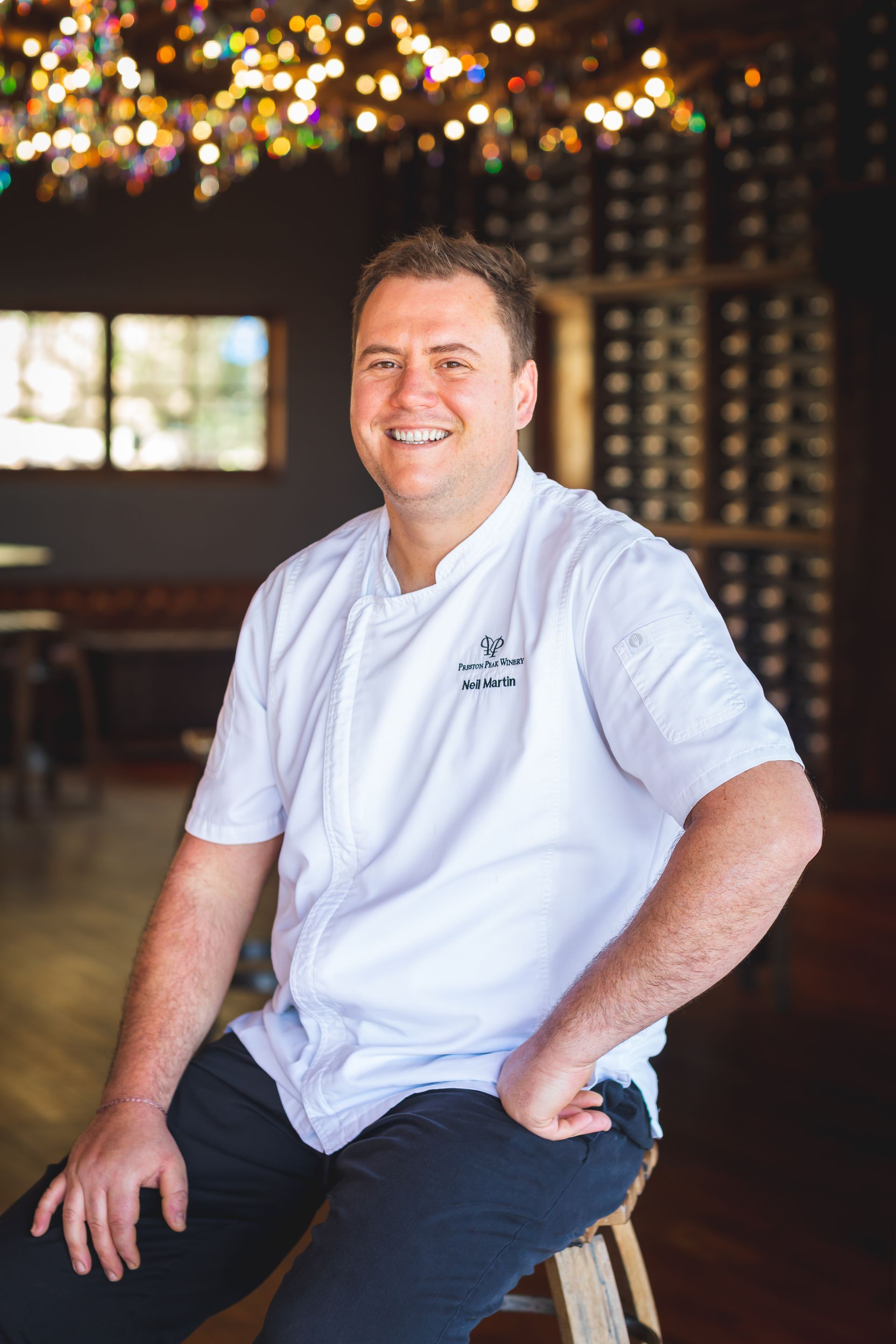 A man in a chef 's uniform is sitting on a stool and smiling.