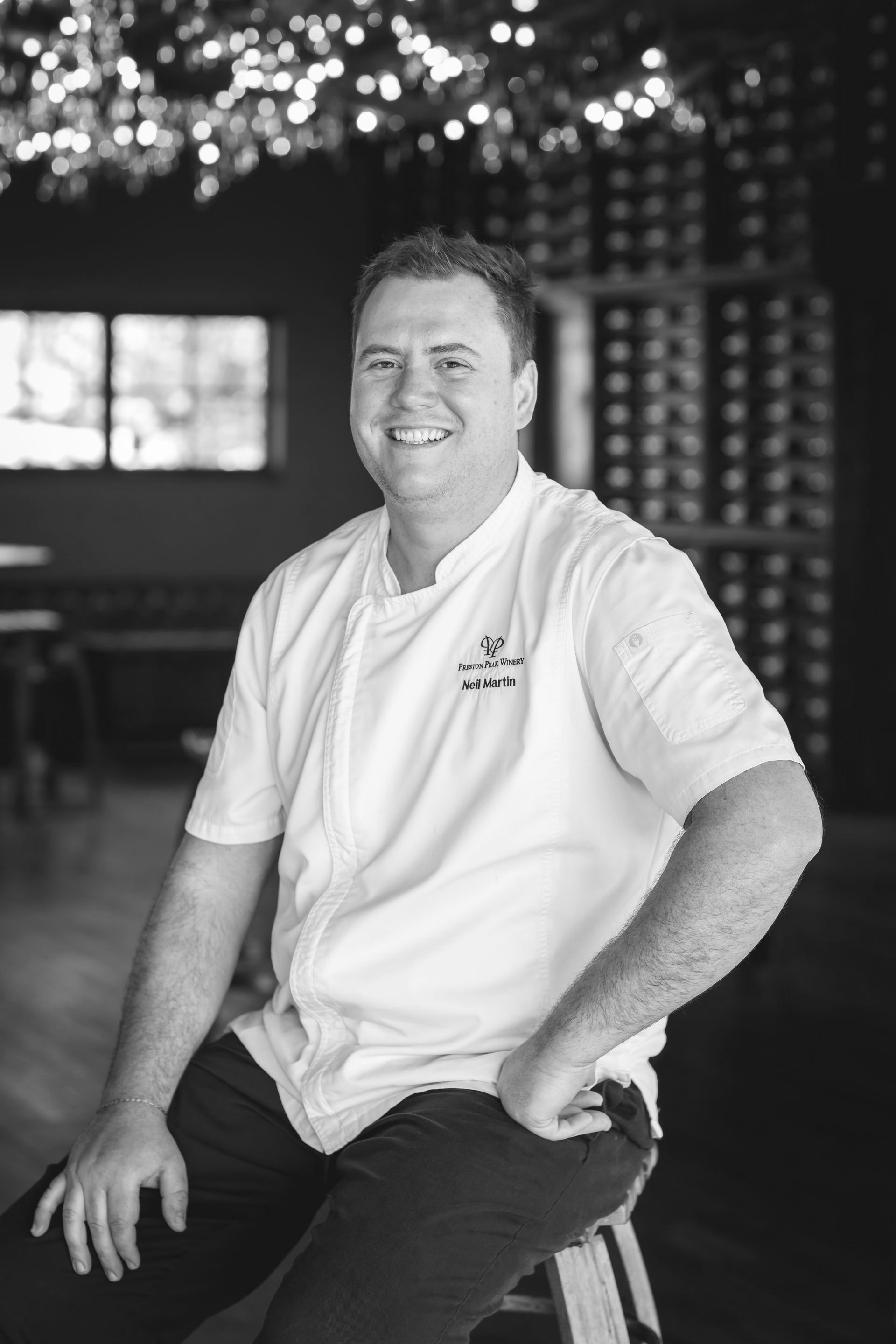 A man in a chef 's uniform is sitting on a stool in a black and white photo.