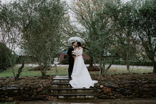 A bride and groom are kissing under an umbrella in the rain.