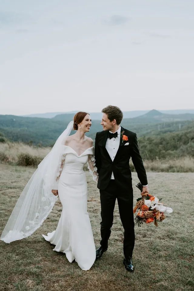 A bride and groom are walking in a field holding hands.