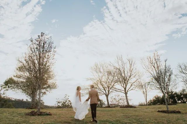 A bride and groom are walking in a field holding hands.
