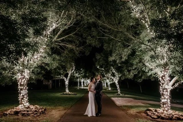 A bride and groom are standing under trees with lights on them at night.