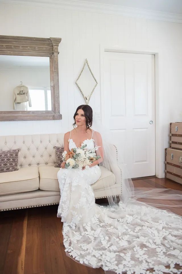 A bride in a long white dress is sitting on a couch holding a bouquet of flowers.