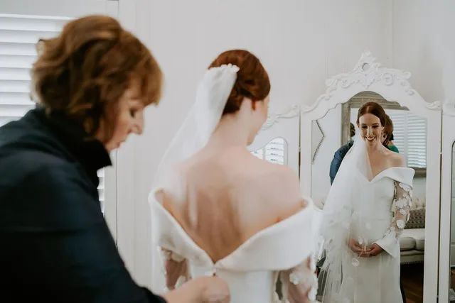 A woman is helping a bride get ready for her wedding in front of a mirror.
