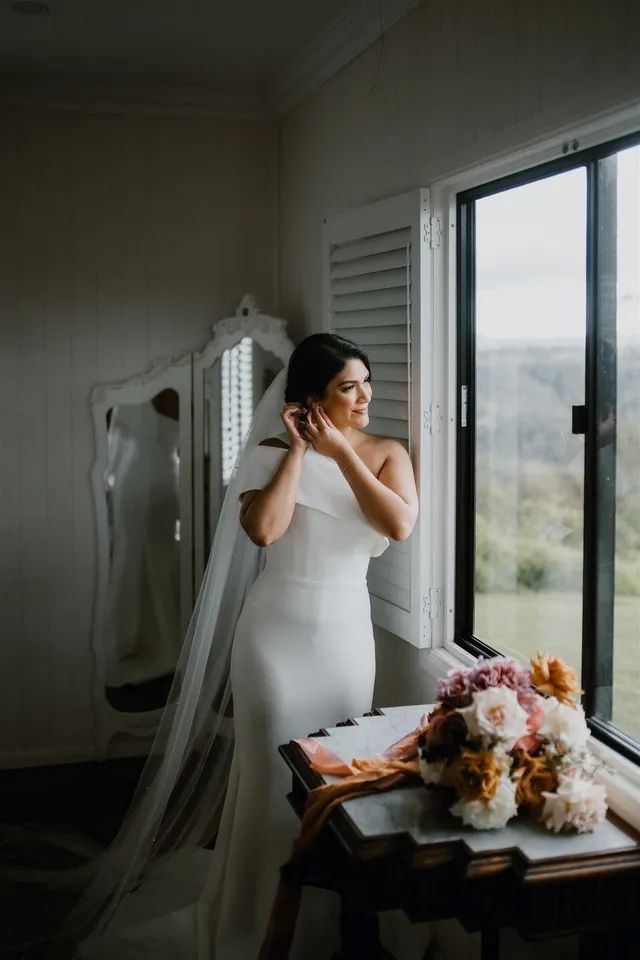 A bride in a wedding dress is standing in front of a window.