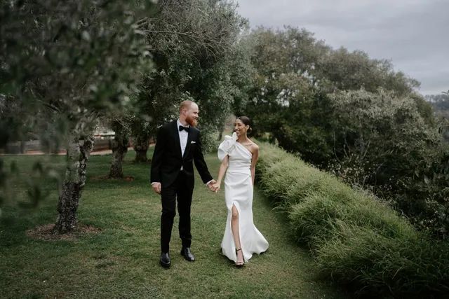 A bride and groom are walking in the grass holding hands.