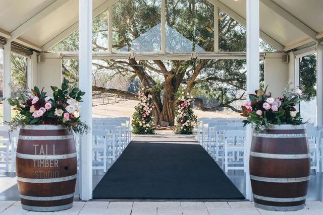 A wedding ceremony is taking place in a gazebo with barrels filled with flowers.