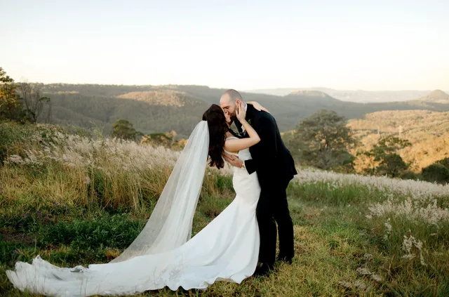 A bride and groom are kissing in a field with mountains in the background.