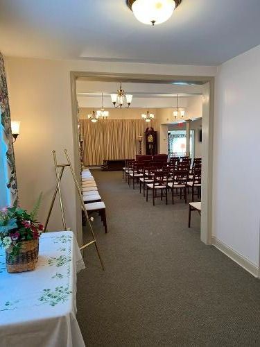 Interior view of a chapel with rows of chairs, a table with flowers, and a doorway leading to the seating area.