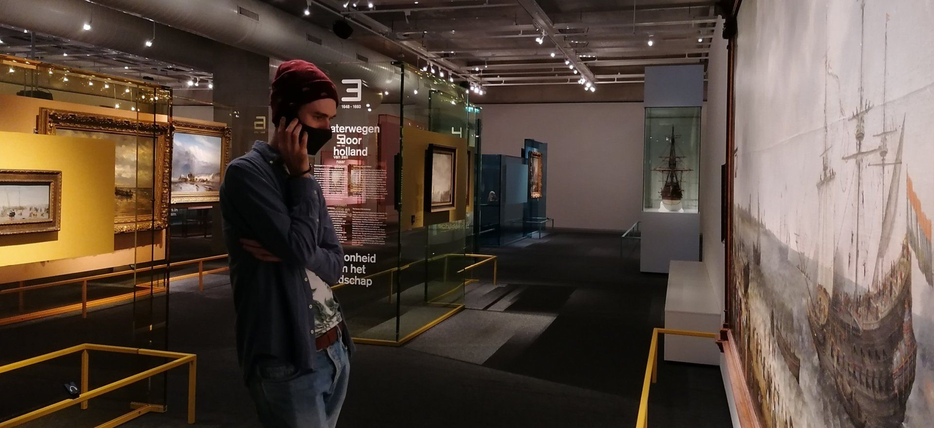 A masked person during the pandemic listening to a Podcatcher in  empty looking museum room with several artworks displayed