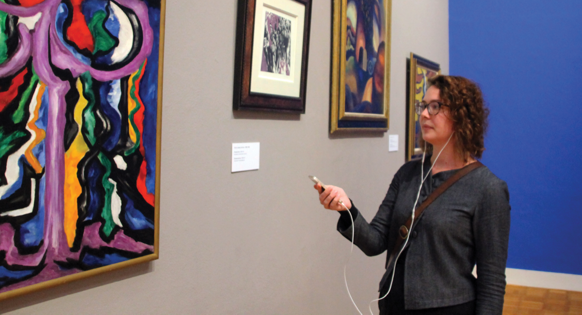 A woman listening to an audio guide whilst looking at a painting on a wall in a museum
