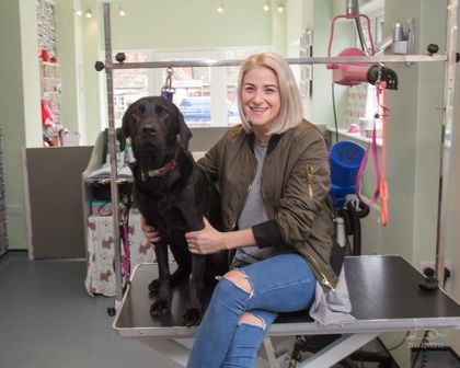 Woman with short blonde hair and black Labrador at a grooming table. She gives a thumbs up.