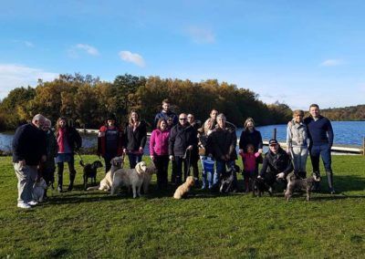 Group of people with dogs posing on grassy bank near water on a sunny day.