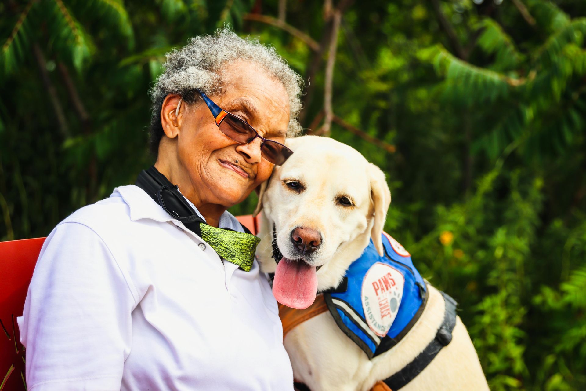 Woman in glasses smiles, hugging a yellow lab wearing a blue vest outdoors.