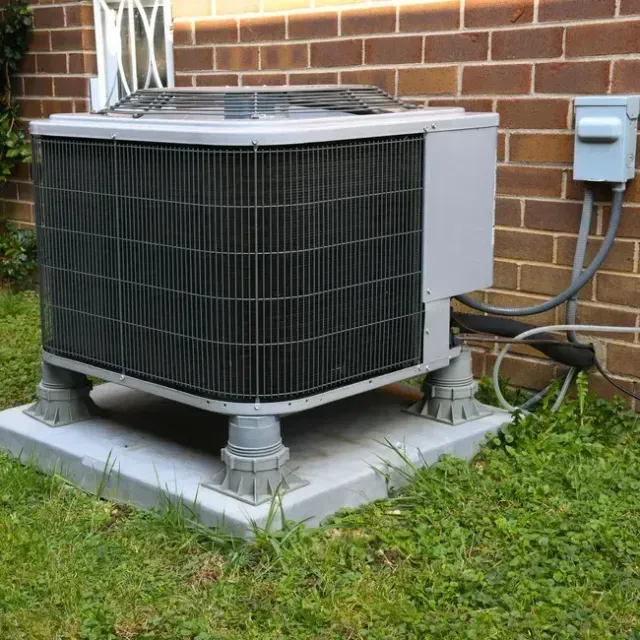 Air conditioning unit on a gray concrete pad with gray plastic supports, set against a brick wall.