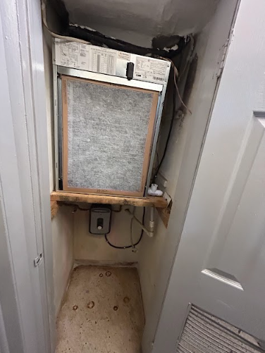 HVAC air filter inside a small, narrow closet. Filter is dusty; wooden shelf and electrical box below.