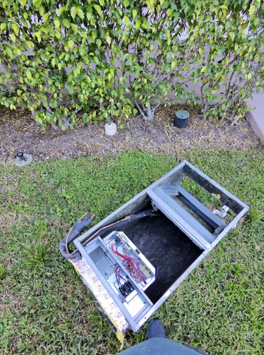 Open air conditioning unit lying on grass, with a hedge background.