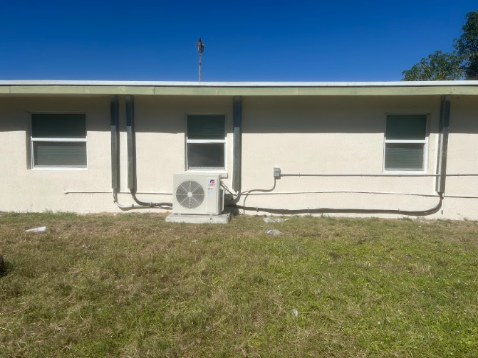 Back exterior of a beige building with windows and an AC unit on grass under a clear blue sky.