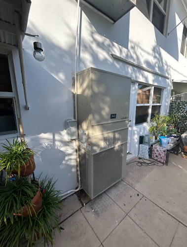 A beige air conditioning unit attached to a white building exterior with windows.