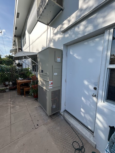 Air conditioning unit next to a white door of a light-colored building. Patio furniture and plants are nearby.