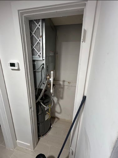 Furnace in a closet, with a white filter, pipes, and a thermostat on the wall.