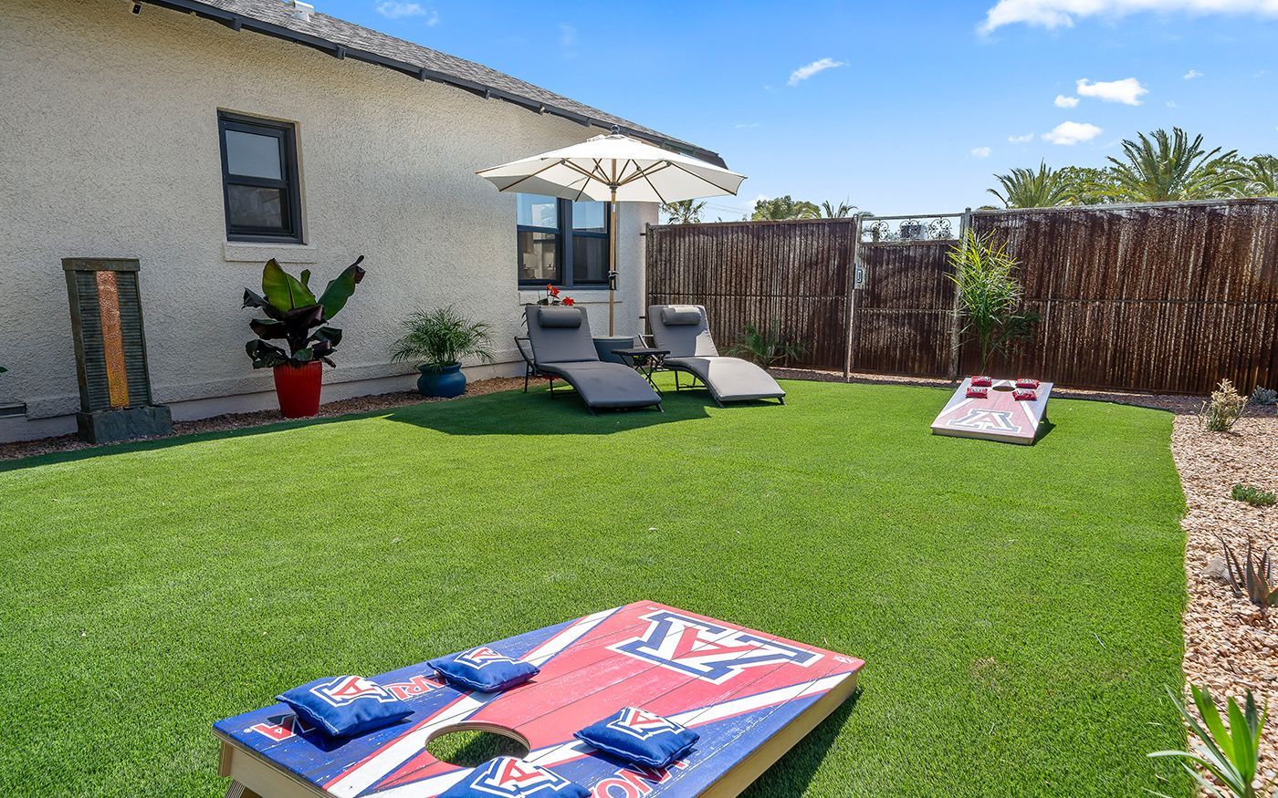 Backyard with artificial grass, lounge chairs under an umbrella, cornhole boards.
