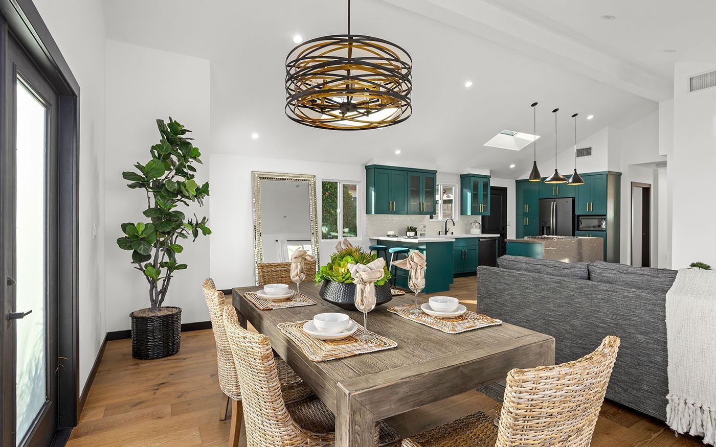 Dining room with wood table, wicker chairs, green kitchen, and large leafy plant.